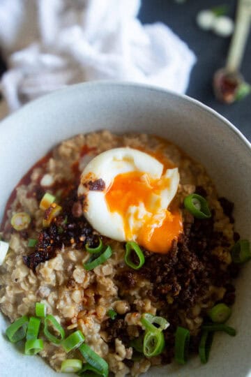 Savory Asian Oatmeal topped with crispy beef, chilli oil, spring onions and poached egg in a white bowl.